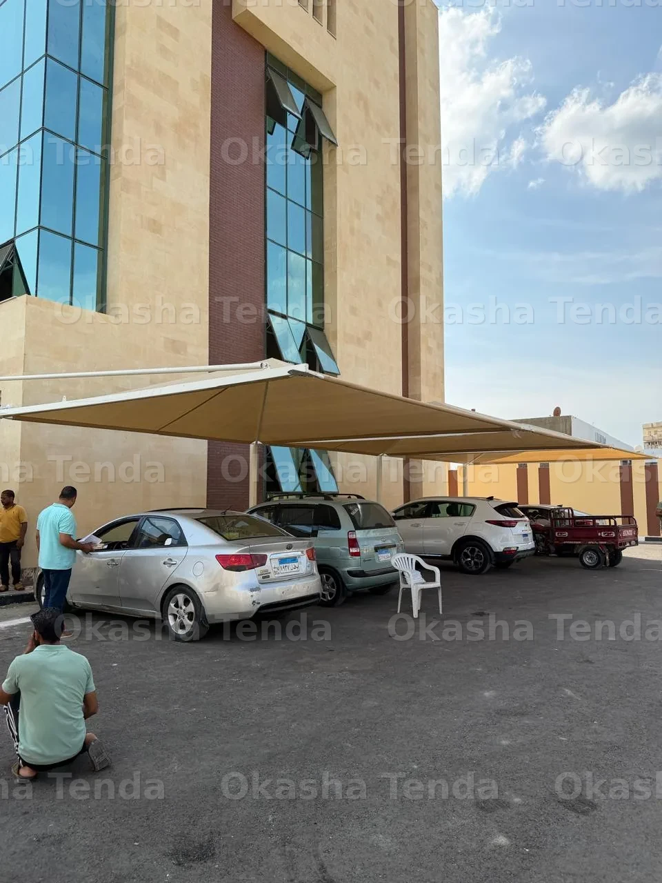 مظلة سيارات مثبتة بقماش مظلة بني تغطي عدة سيارات بجانب مبنى عقاري.